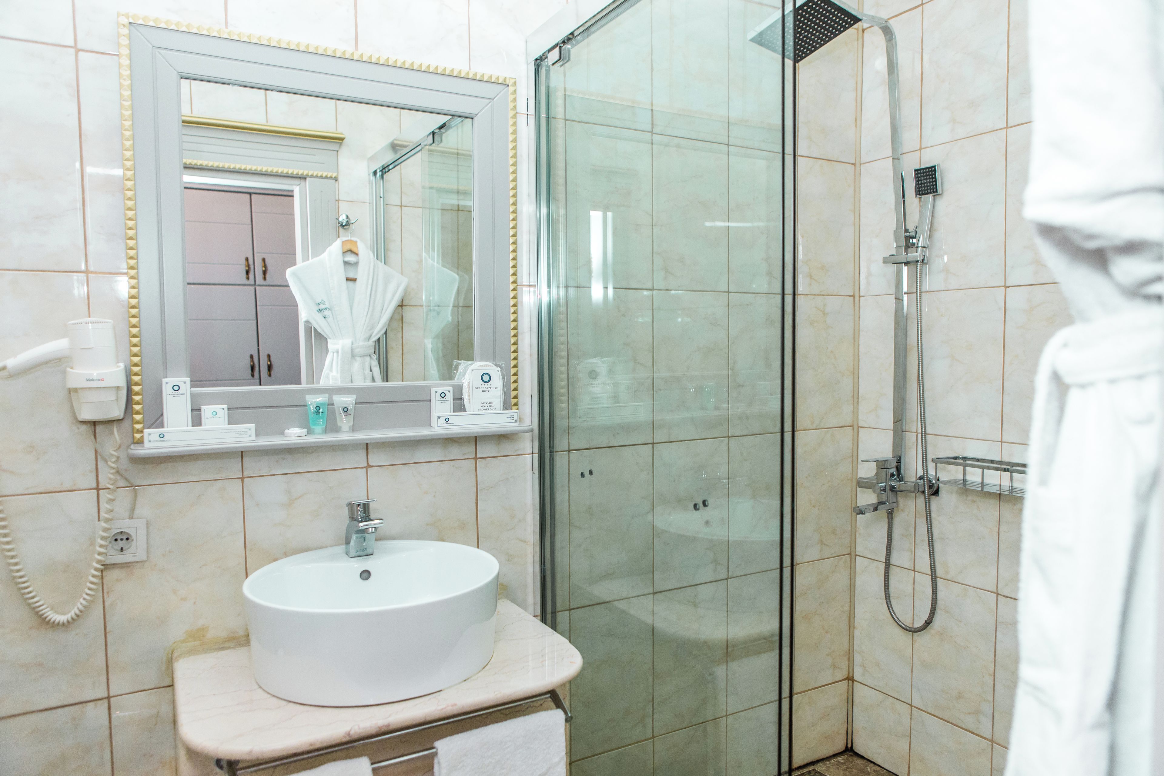 A bathroom with a glass-enclosed shower, a white sink, a mirror with a gold frame, and a towel rack.