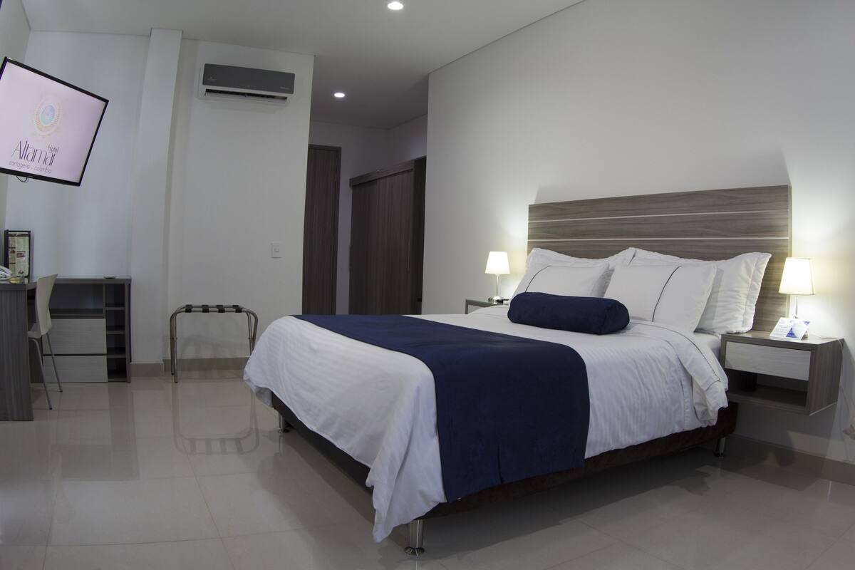 A modern hotel room with a large bed, bedside tables with lamps, a desk with a chair, and a TV mounted on the wall.