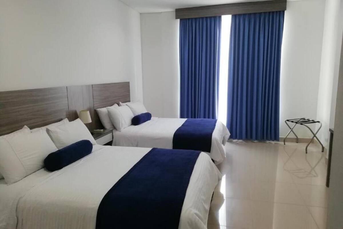 A hotel room with two beds, a wooden headboard, a window with blue curtains, and a small table with a drying rack.