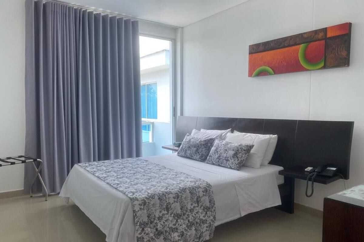 A hotel room with a large bed, a bedside table with a phone, a window with curtains, and a wall art piece.