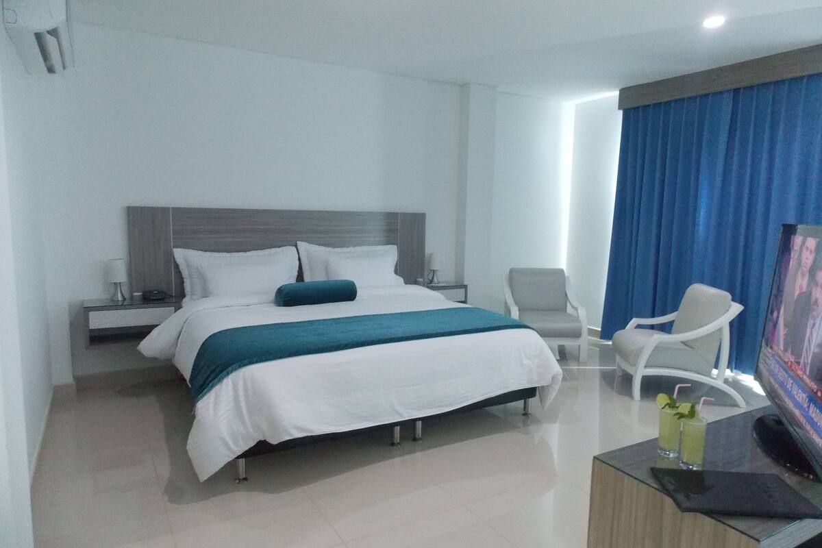 A modern hotel room with a large bed, a desk with a flat-screen TV, a chair, and a small table with a drink.
