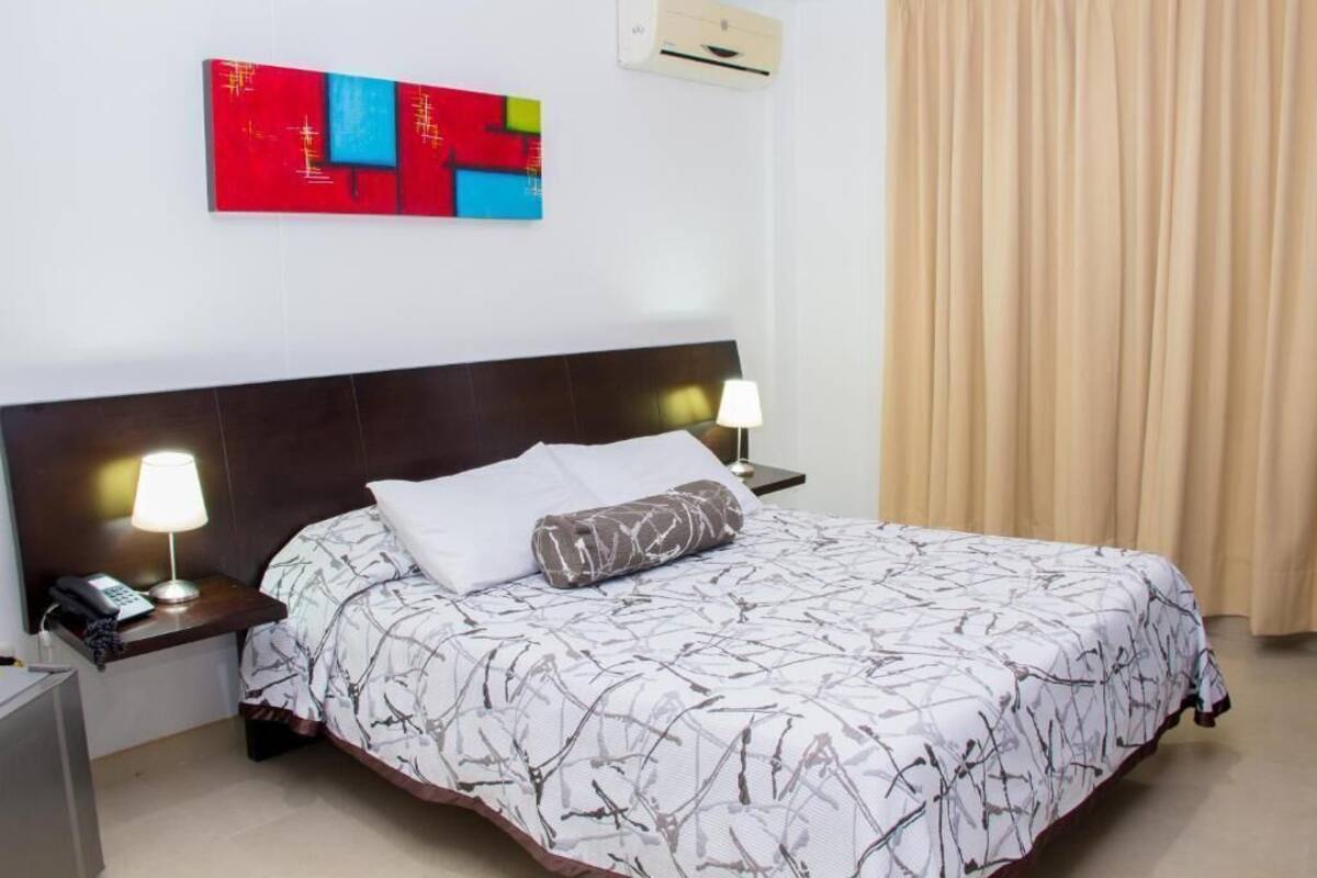 A hotel room with a bed, nightstands, a phone, a lamp, an air conditioner, and a colorful abstract painting on the wall.