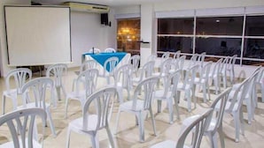 A conference room with rows of white plastic chairs, a blue table, and a projector screen. - Hotel Altamar (Cartagena)