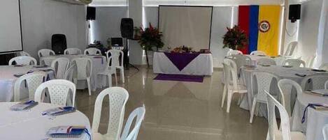 Un salón de conferencias con sillas y mesas de plástico blanco, un escenario con un atril y una bandera en la pared.