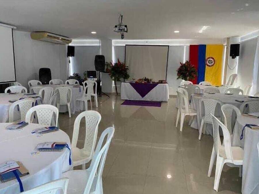 Un salón de conferencias con sillas y mesas de plástico blanco, un escenario con un atril y una bandera en la pared.