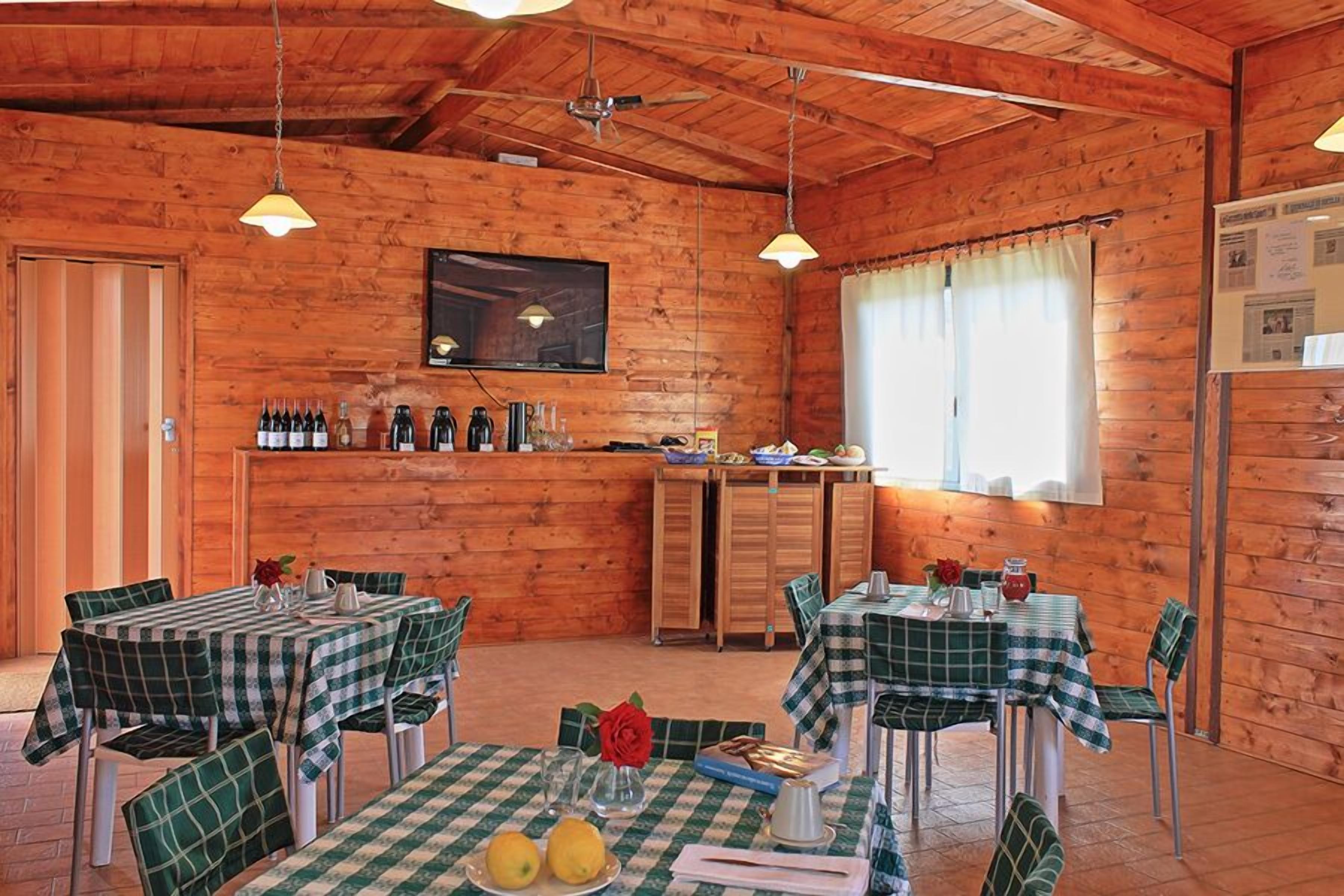 A wooden cabin with multiple dining tables covered with checkered tablecloths, a bar area with bottles and glasses, and a television mounted on the wall.