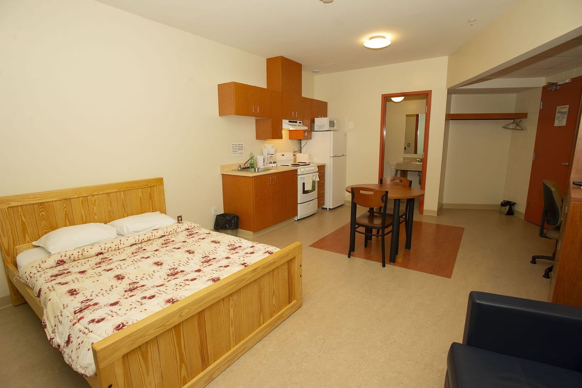 Un dormitorio con una cama de madera, una pequeña kitchenette y un baño visible a través de una puerta abierta.