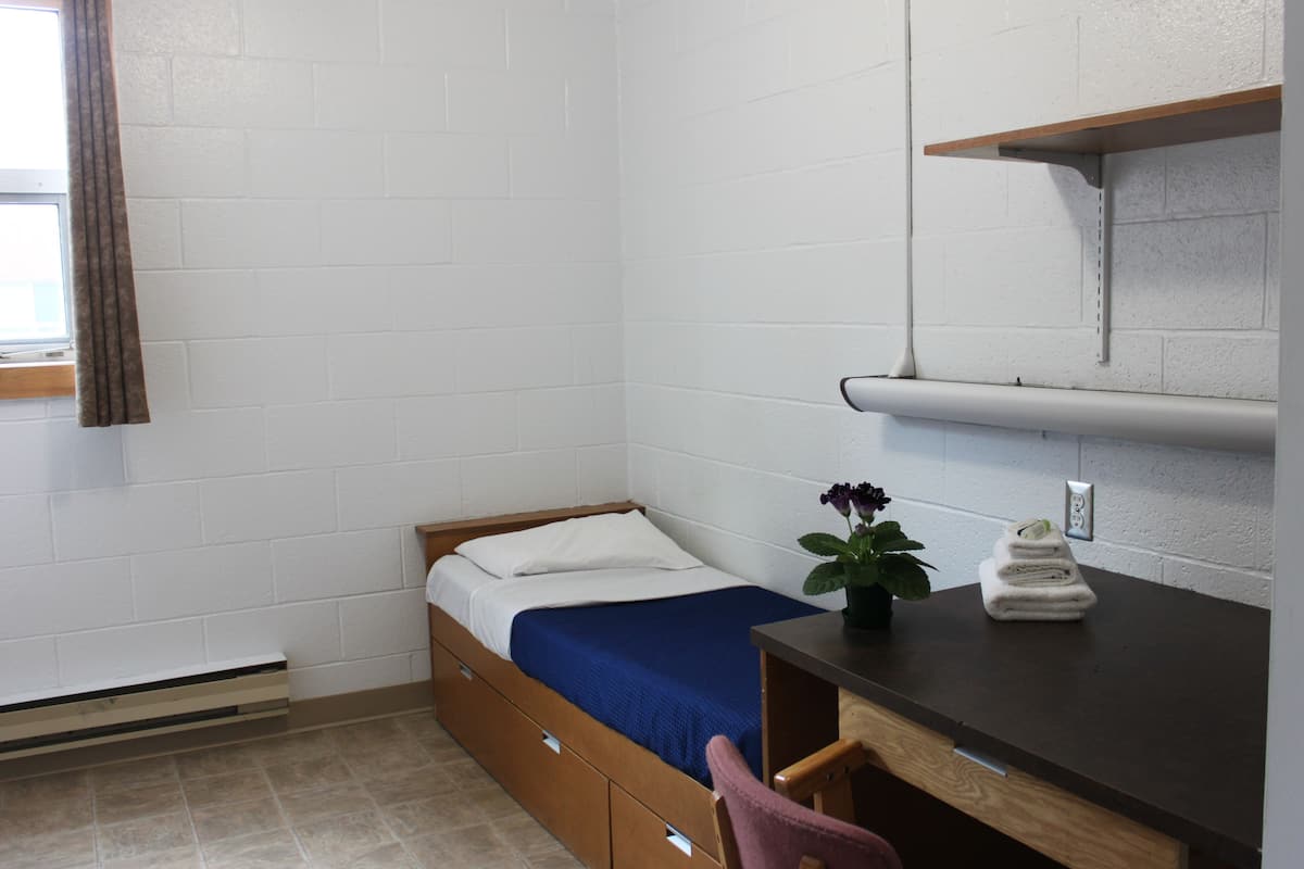 Una habitación pequeña y limpia con una cama individual, un escritorio y una silla. Hay una ventana con cortinas, una planta en el escritorio y una toalla sobre la cama.