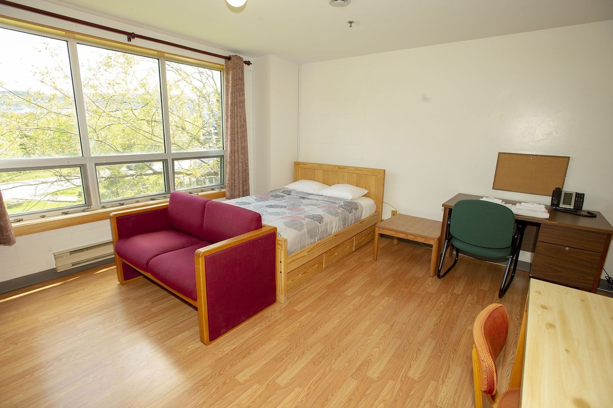 Una habitación con cama, escritorio, silla y ventana con vistas a árboles.