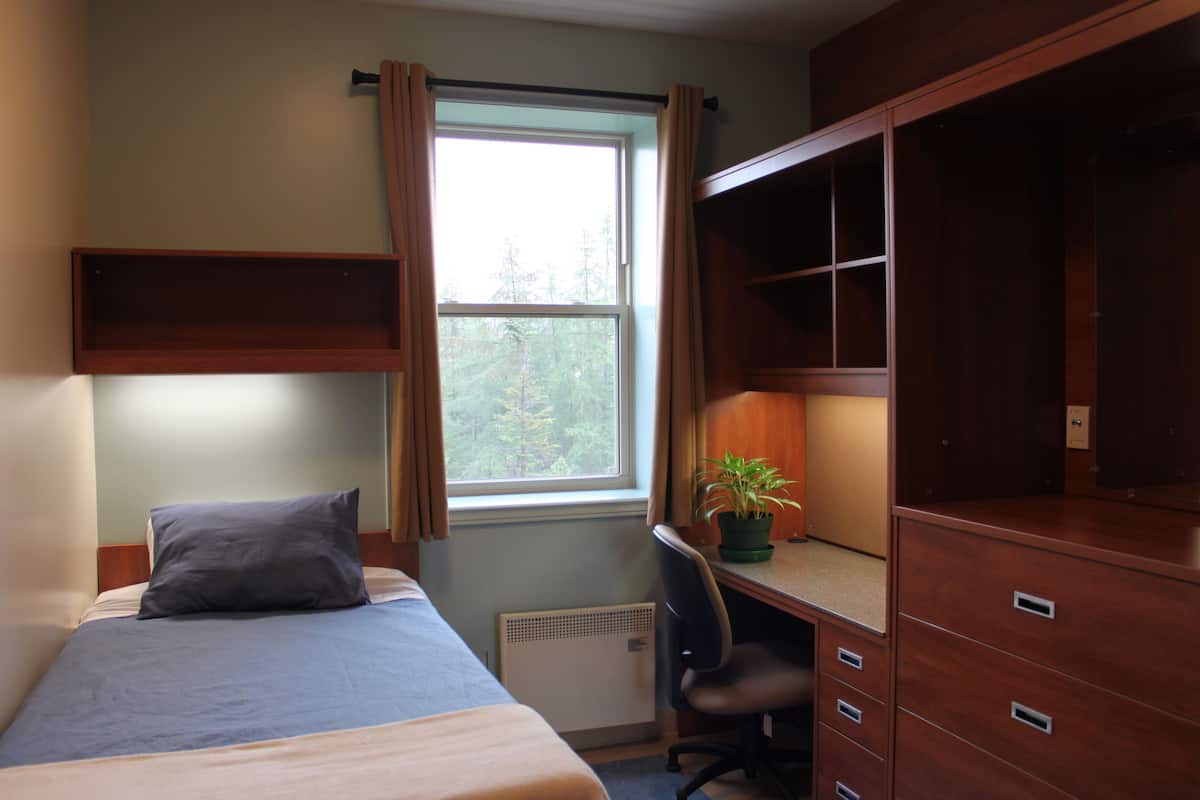 Un dormitorio con cama, escritorio, silla, radiador y ventana con cortinas.