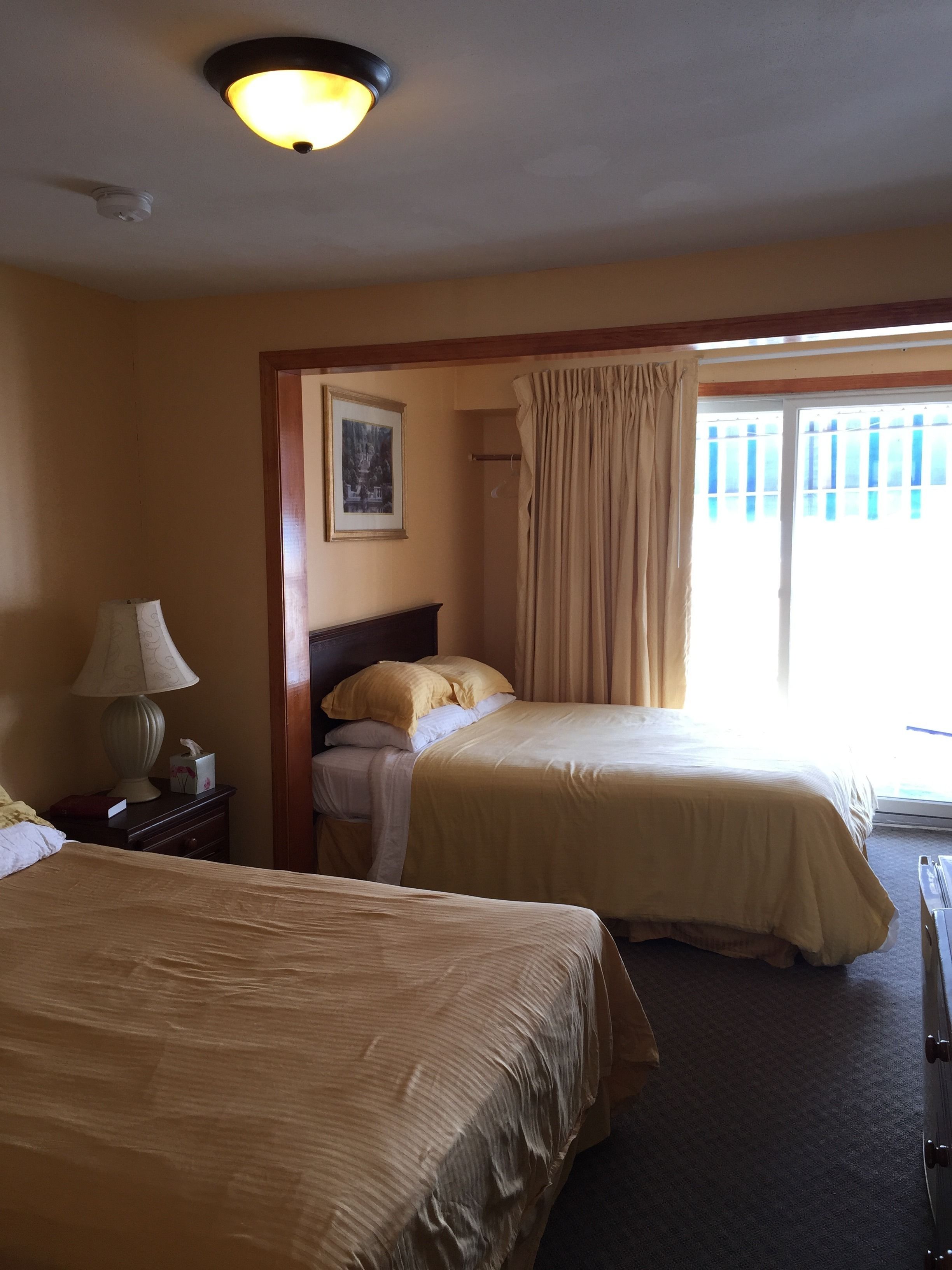 Habitación de hotel con dos camas, una mesita de noche con lámpara, un cuadro en la pared y una ventana con cortinas.