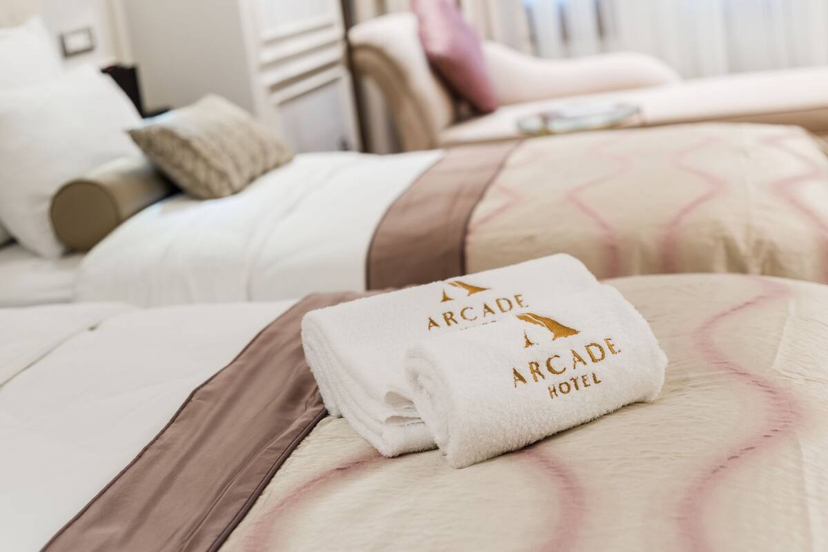 Cama bem arrumada com uma toalha branca dobrada, exibindo o texto "Arcade Hotel".