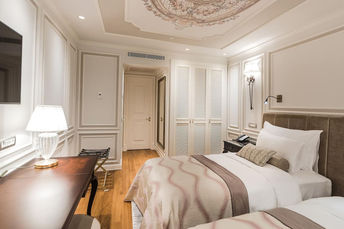 Quarto de hotel com mesa de jantar de madeira, cama com colcha riscada em bege e marrom, mesa de cabeceira com abajur e teto com uma moldura decorativa.