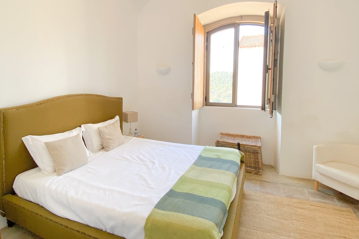 A bedroom with a bed, a window, a bedside table, and a chair.