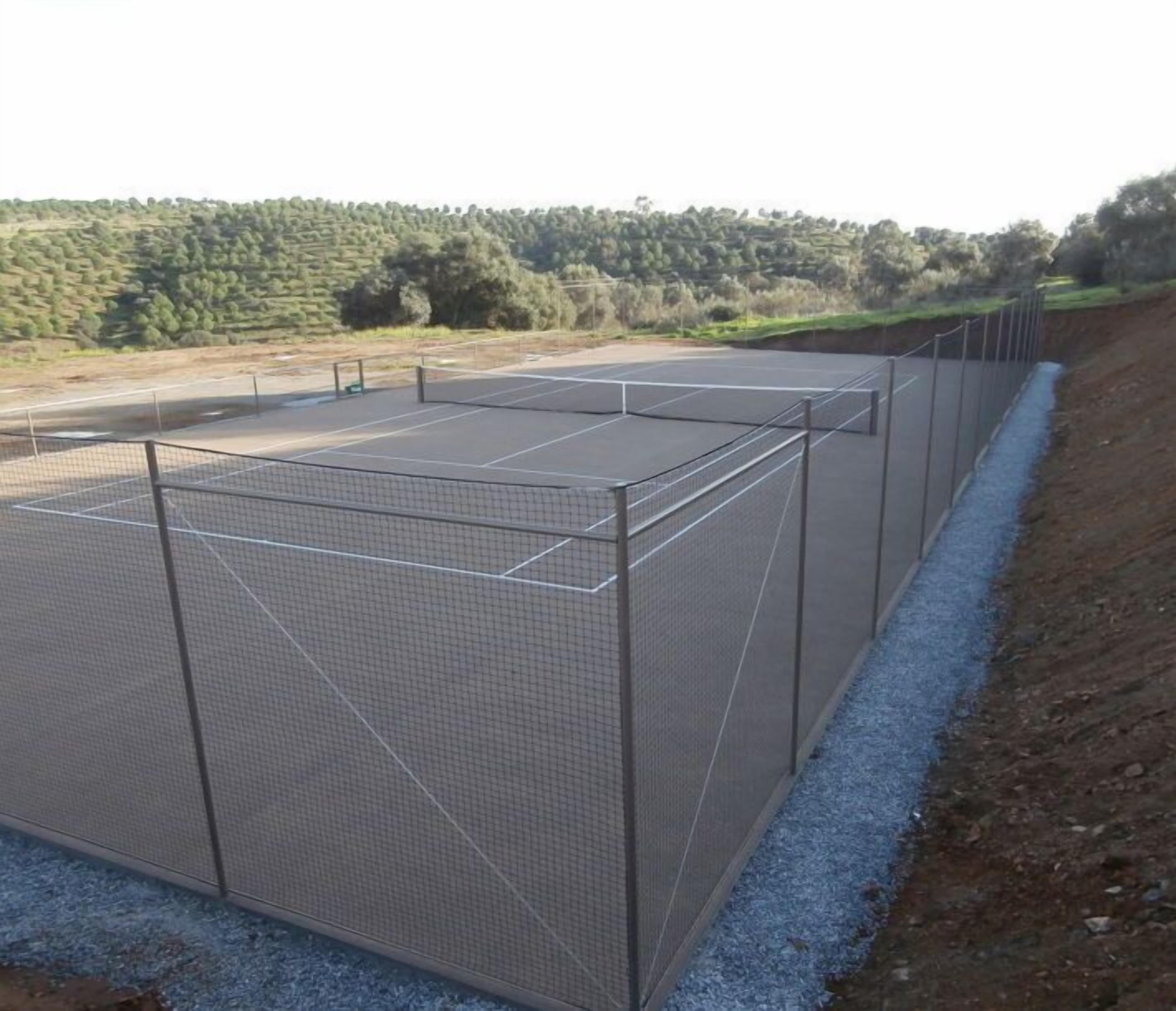 Una cancha de tenis con red y valla perimetral, rodeada por un sendero de grava y un terraplén de tierra.