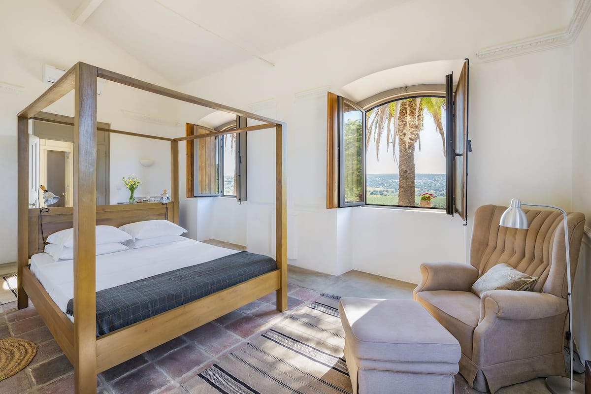 A four-poster bed with a view of palm trees and the sea through a window.