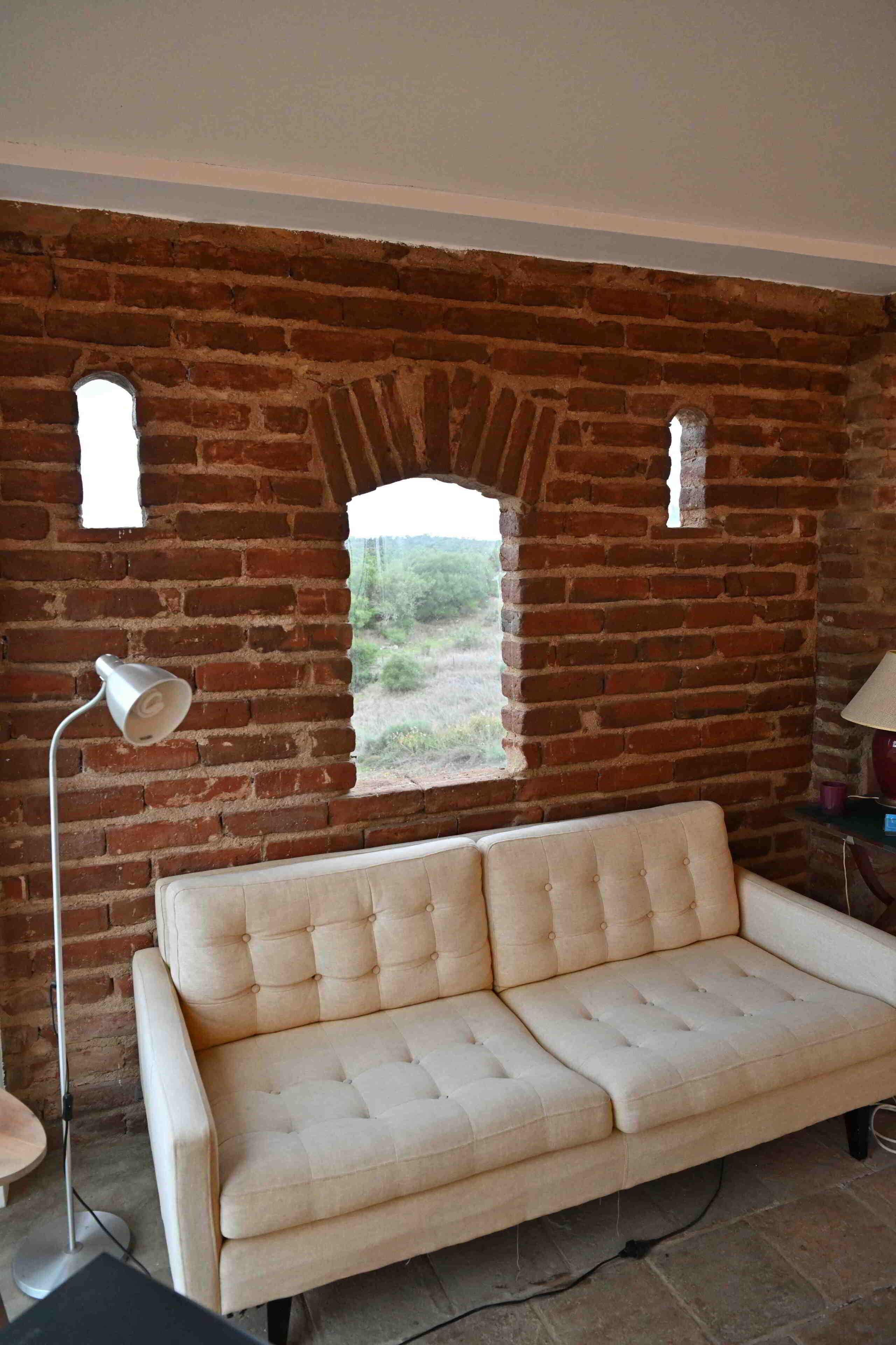 Una habitación con pared de ladrillo, ventana, sofá color beige, lámpara de pie y una mesa.