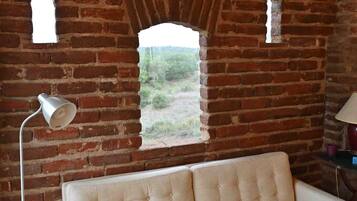 Una habitación con pared de ladrillo, ventana, sofá color beige, lámpara de pie y una mesa.