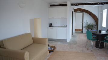 Un salon avec un canapé beige, une table à manger avec des chaises, un coin cuisine avec des meubles blancs et une arche en briques.