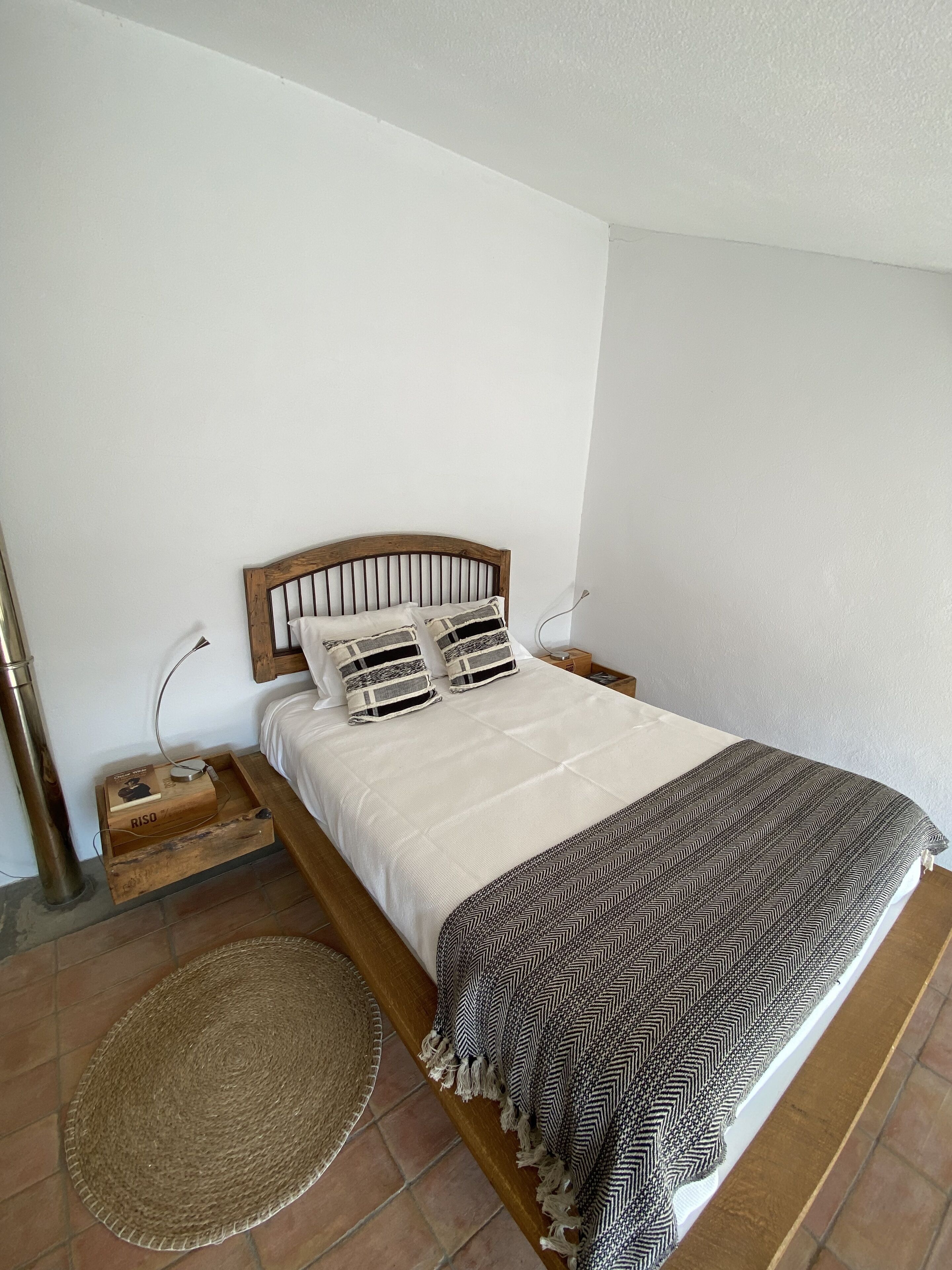 A neatly made bed with a wooden headboard, a patterned throw blanket, and a woven rug on a tiled floor.