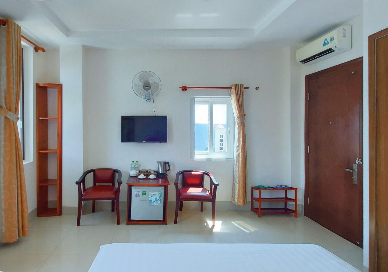 Een hotelkamer met een bed, twee stoelen, een klein tafeltje met een waterkoker, een televisie, een raam met gordijnen, een airconditioner en een deur.