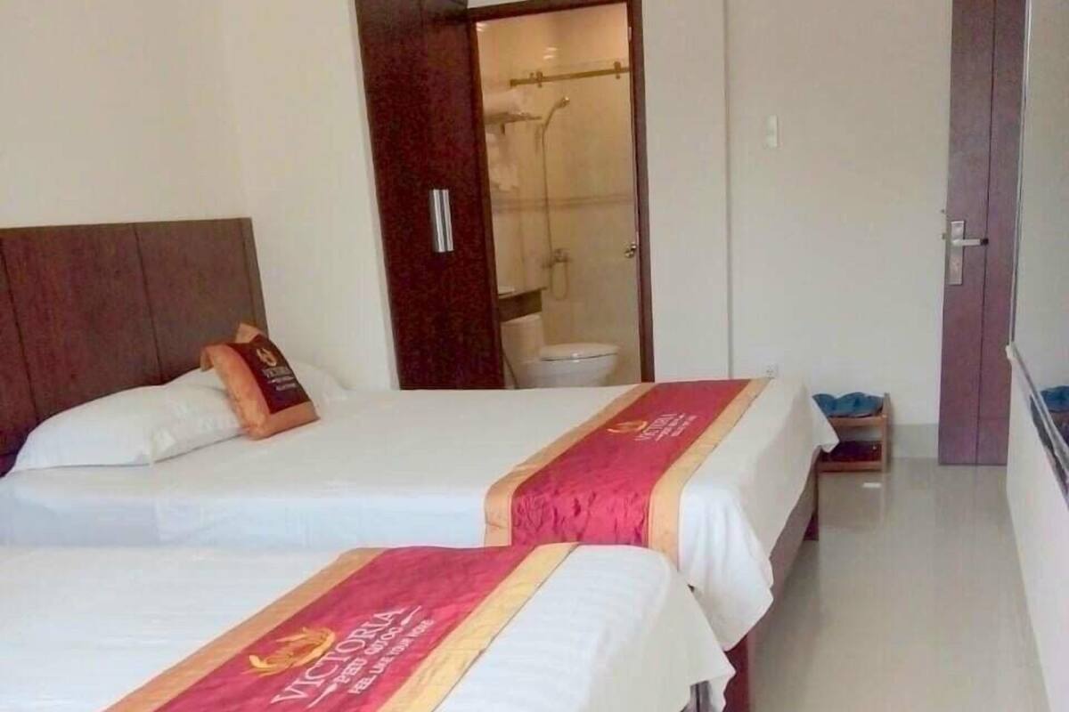 A hotel room with two beds, each with a red and white bedspread, a wooden headboard, and a small bedside table with a blue item on it.