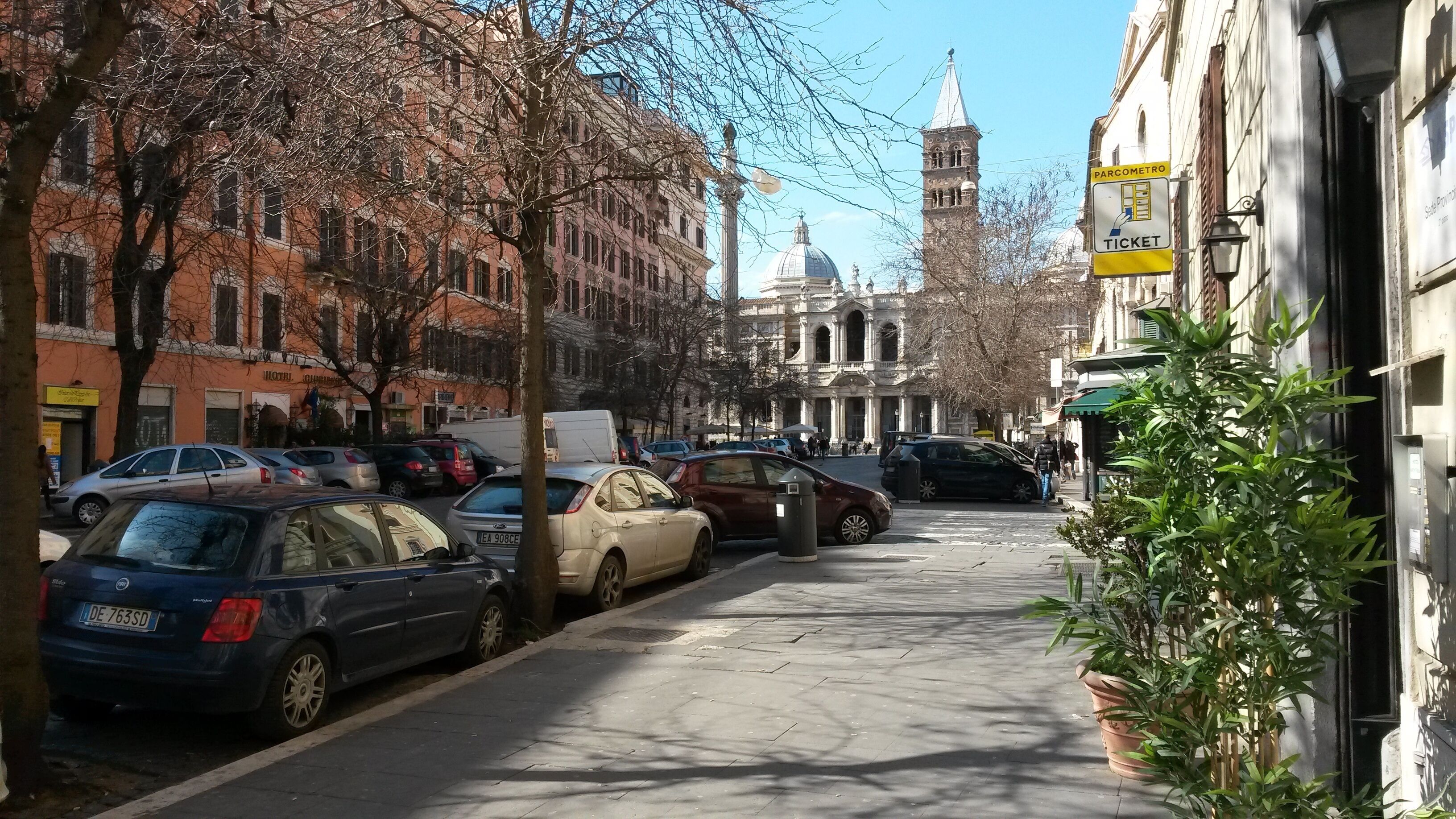 Una calle bordeada de coches aparcados y edificios, incluyendo una iglesia destacada con una cúpula y un campanario.