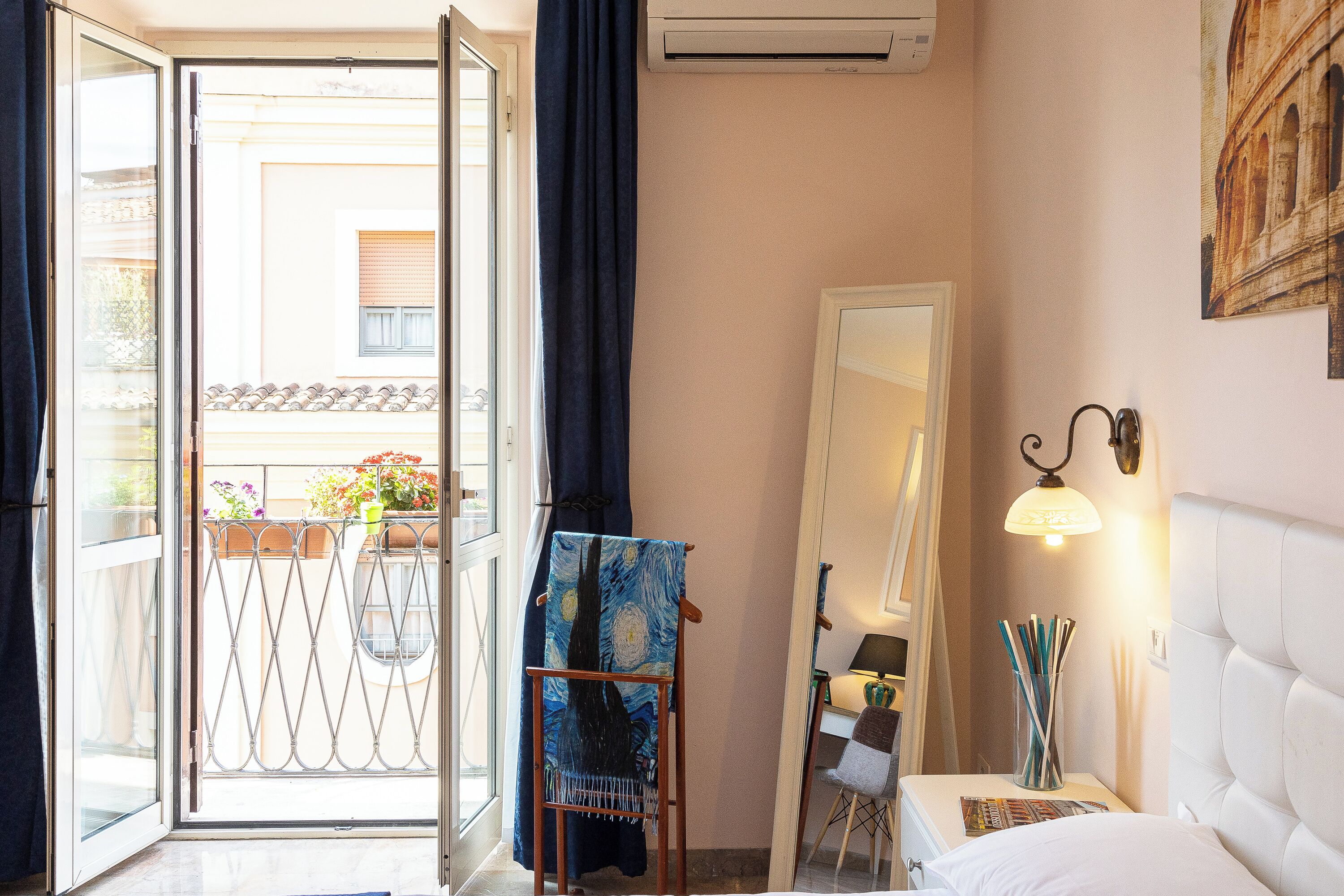 A hotel room with a balcony, a bed with a white headboard, a wall-mounted lamp, and a painting of the Eiffel Tower.
