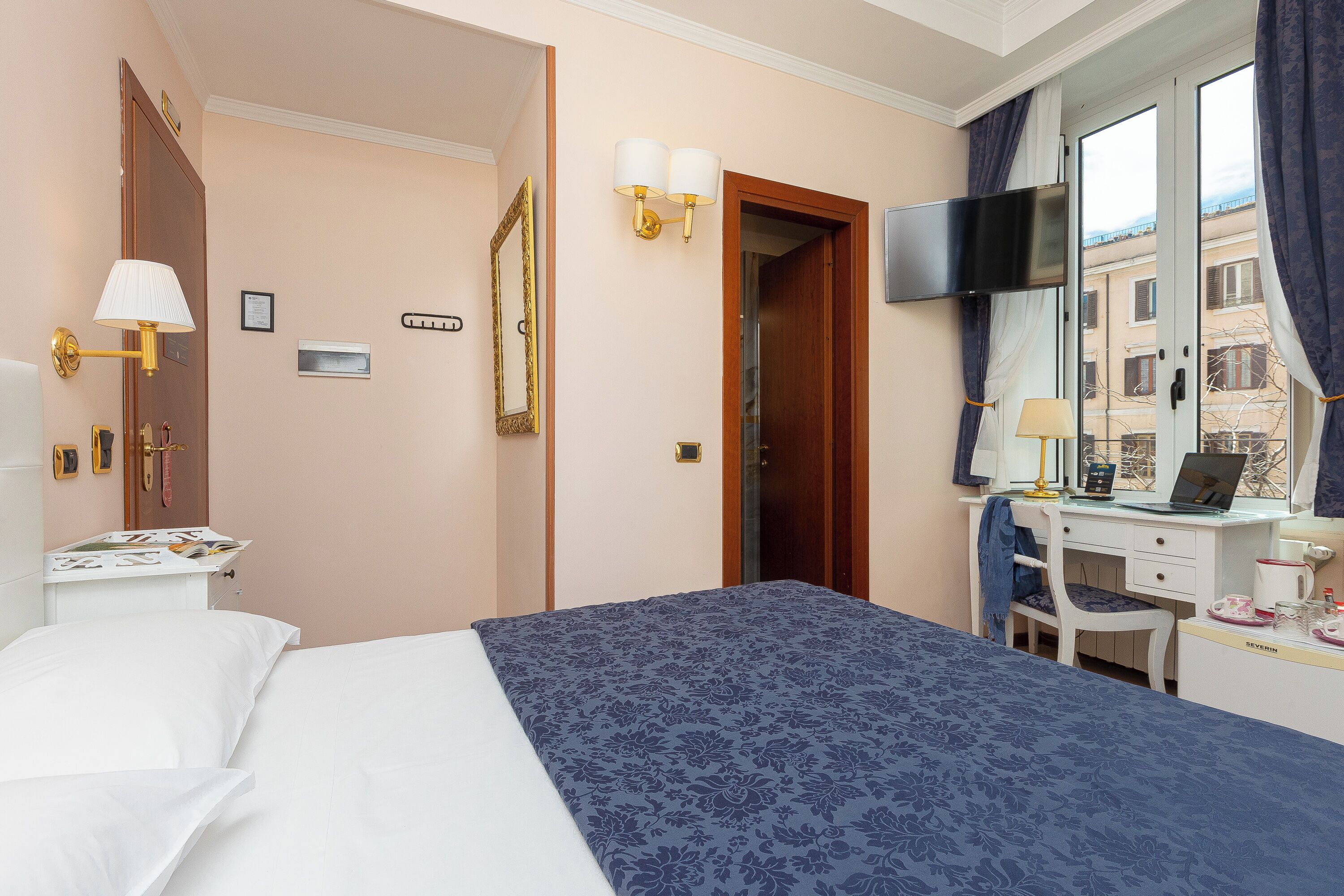 A hotel room with a bed, a desk with a laptop, a TV, and a window with curtains.