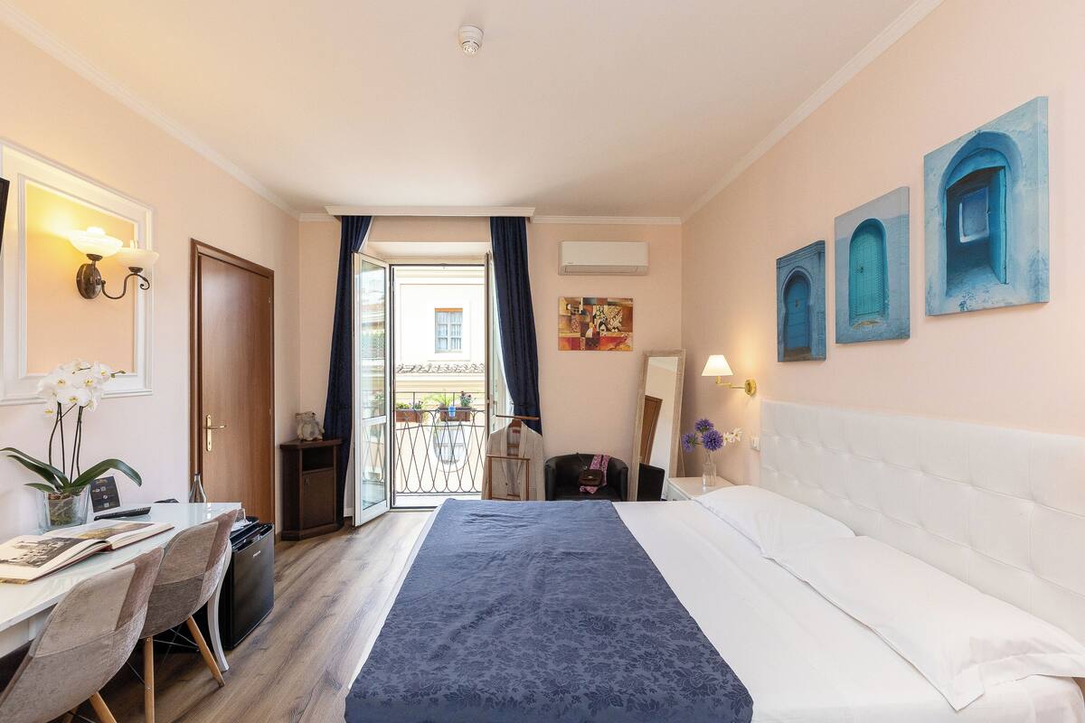 Une chambre d’hôtel avec un lit, un bureau et une chaise, une petite table, une plante en pot et une vue sur l’extérieur.