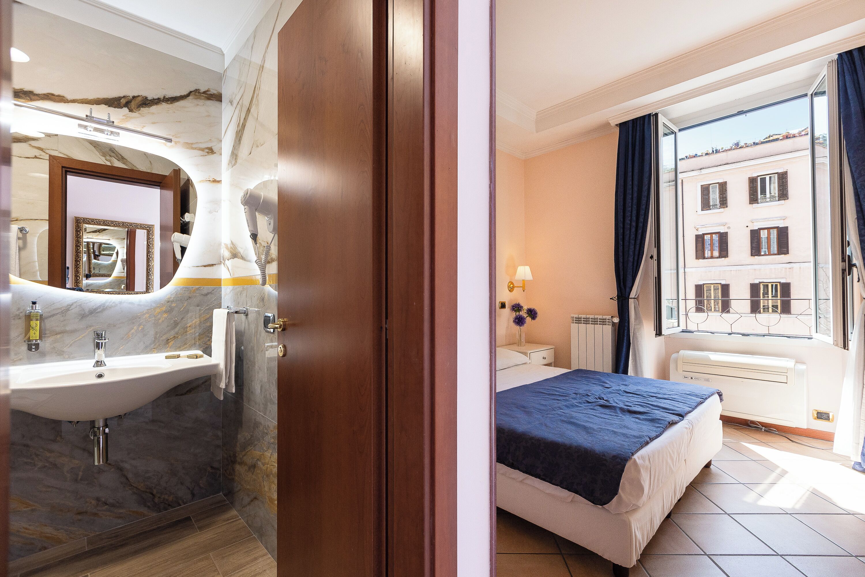 A hotel room with a marble bathroom, a bed with a blue bedspread, and a view of a building through the window.