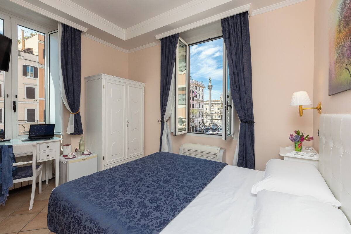 Une chambre d’hôtel avec un lit, un bureau avec un ordinateur portable, une armoire et une vue sur la ville par la fenêtre.