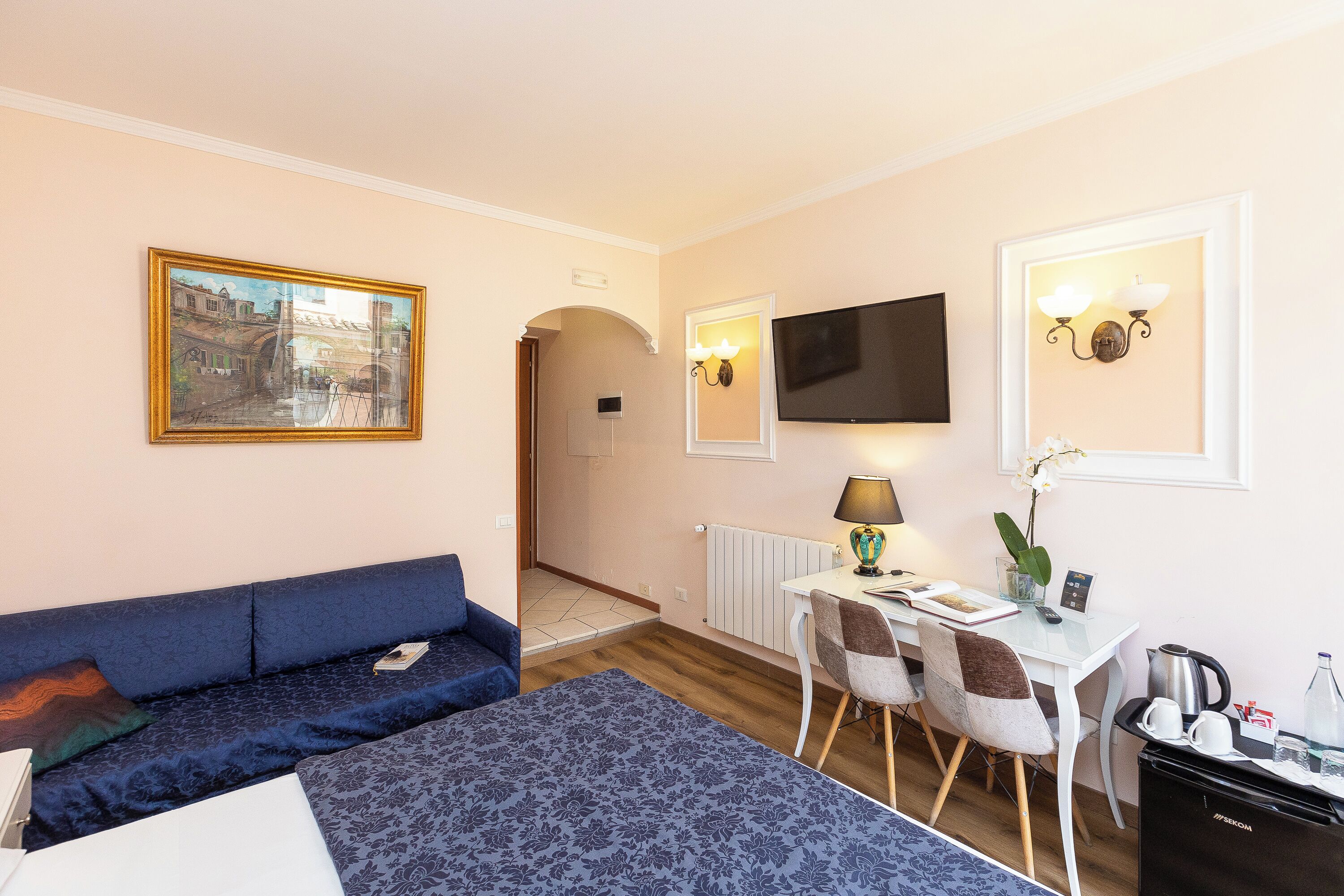 A hotel room with a bed, a sofa, a desk with a chair, a television, and a painting on the wall.