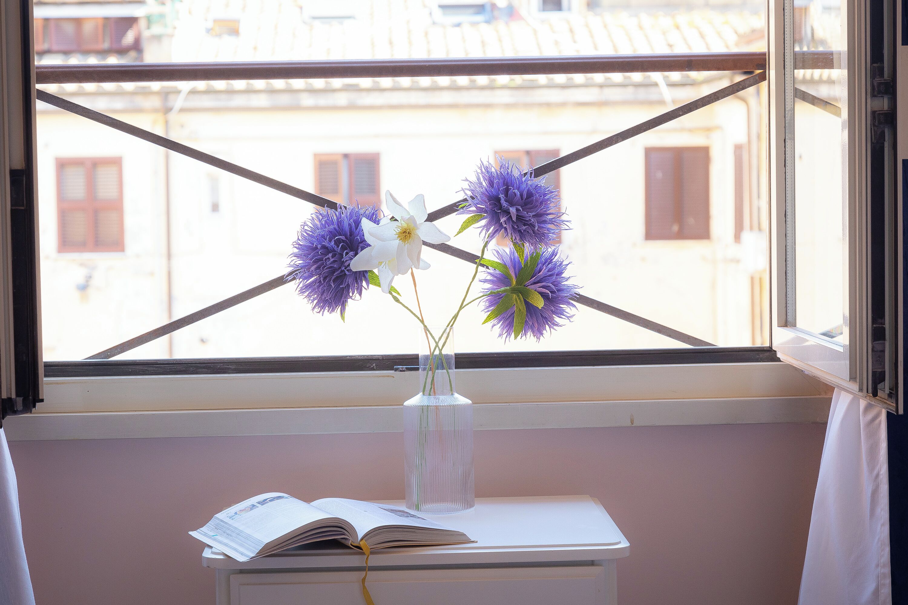 Una ventana con vistas a edificios, un florero con flores y un libro abierto sobre una mesa.