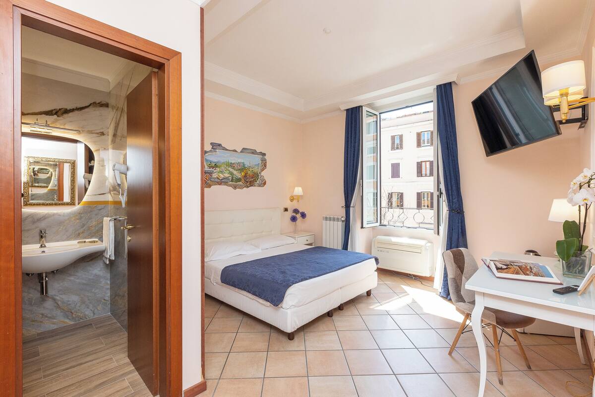 Une chambre d’hôtel avec un lit, un bureau, une télévision, une fenêtre avec des rideaux, une salle de bain avec un lavabo et un tableau au mur.