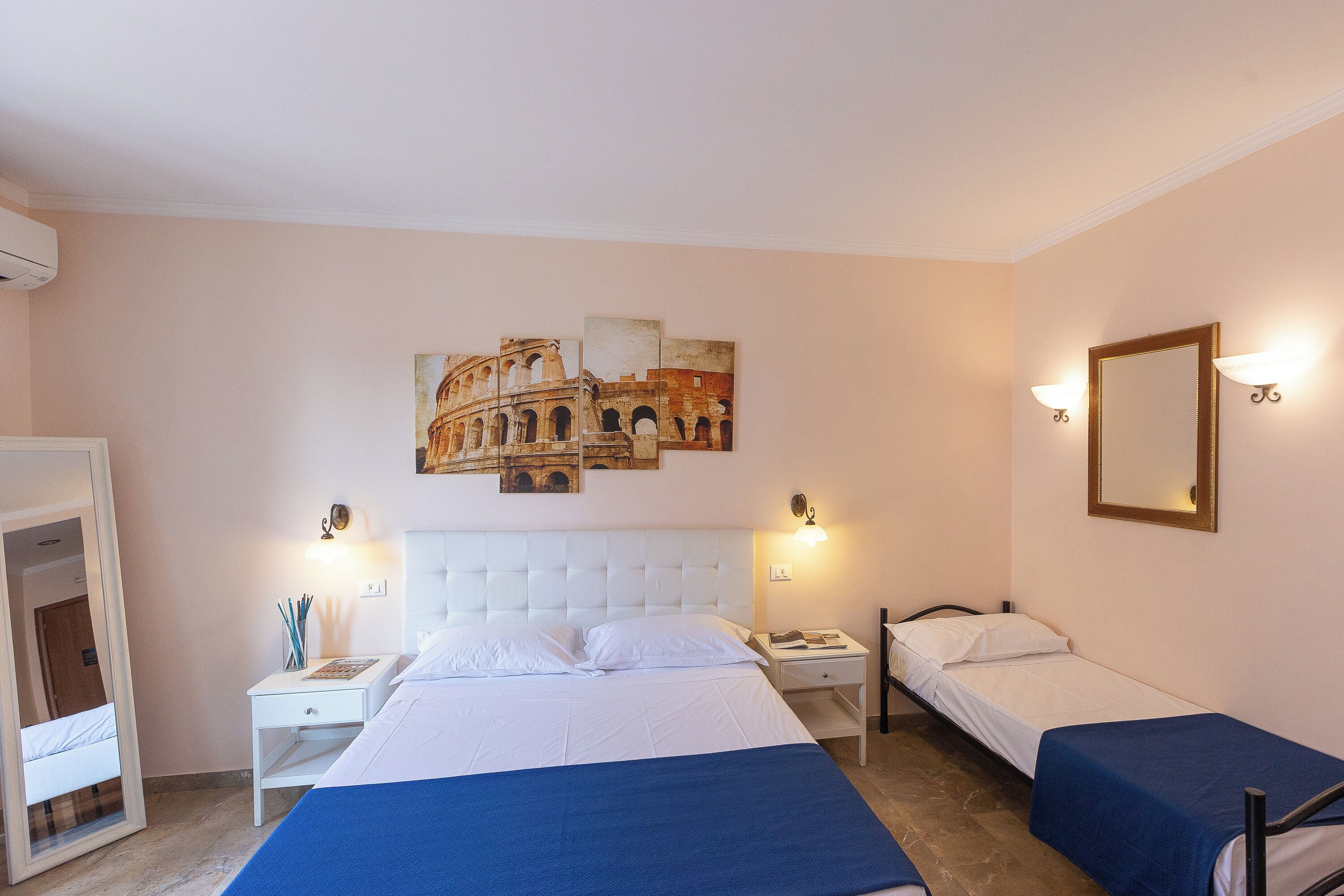 A hotel room with a large bed, two bedside tables, a nightstand, a mirror, and a wall-mounted picture of an ancient structure.