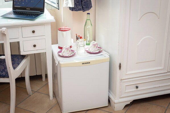 Un refrigerador petit amb exterior blanc i una etiqueta de marca, un ordinador portàtil sobre una taula blanca, una cadira amb coixí floral i un armari amb nansa corbada.
