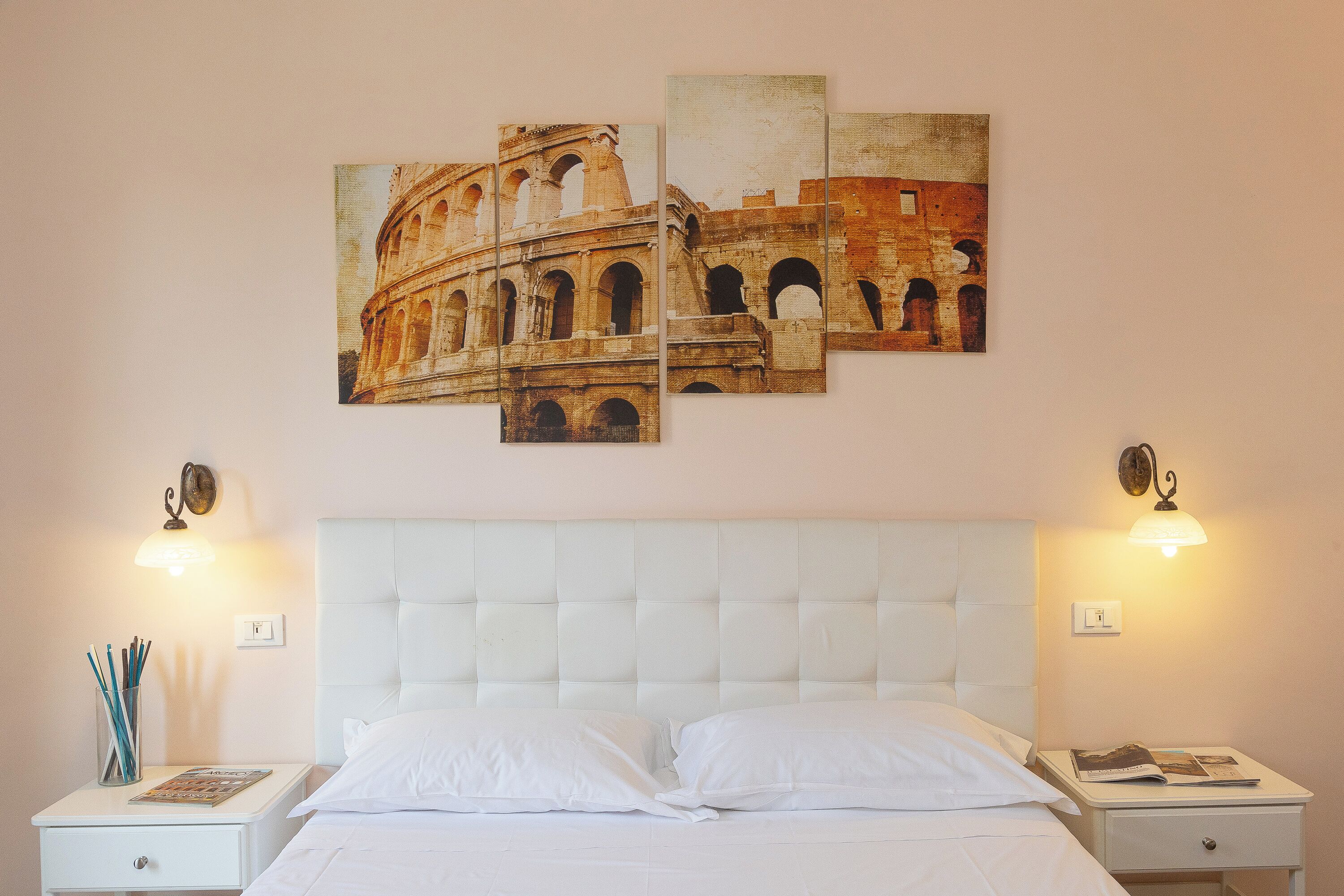 A hotel room with a white bed, two nightstands, a wall-mounted artwork of the Colosseum, and wall sconces.
