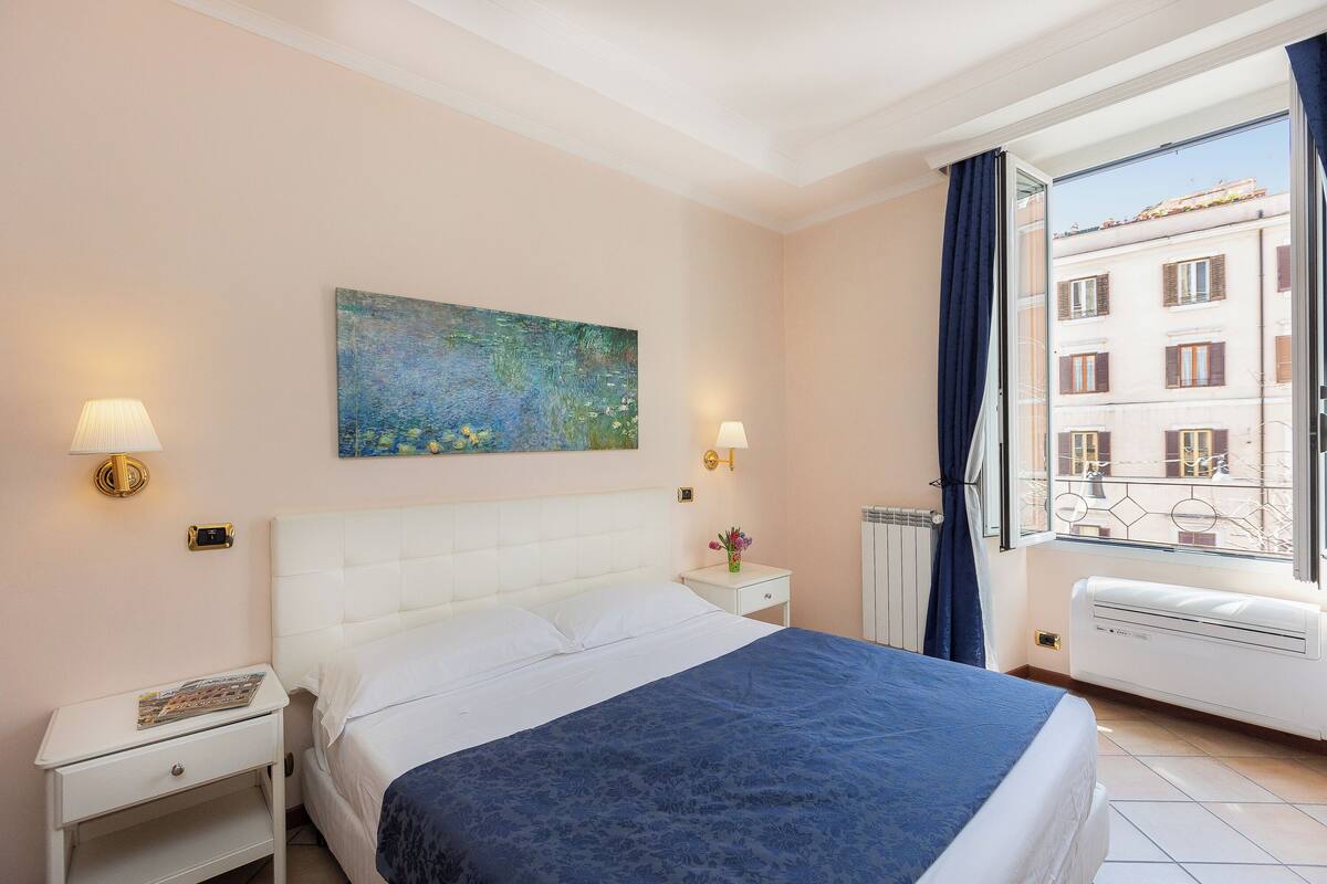 Une chambre d’hôtel avec un lit, des tables de chevet, un tableau, une fenêtre donnant sur des immeubles et un radiateur.