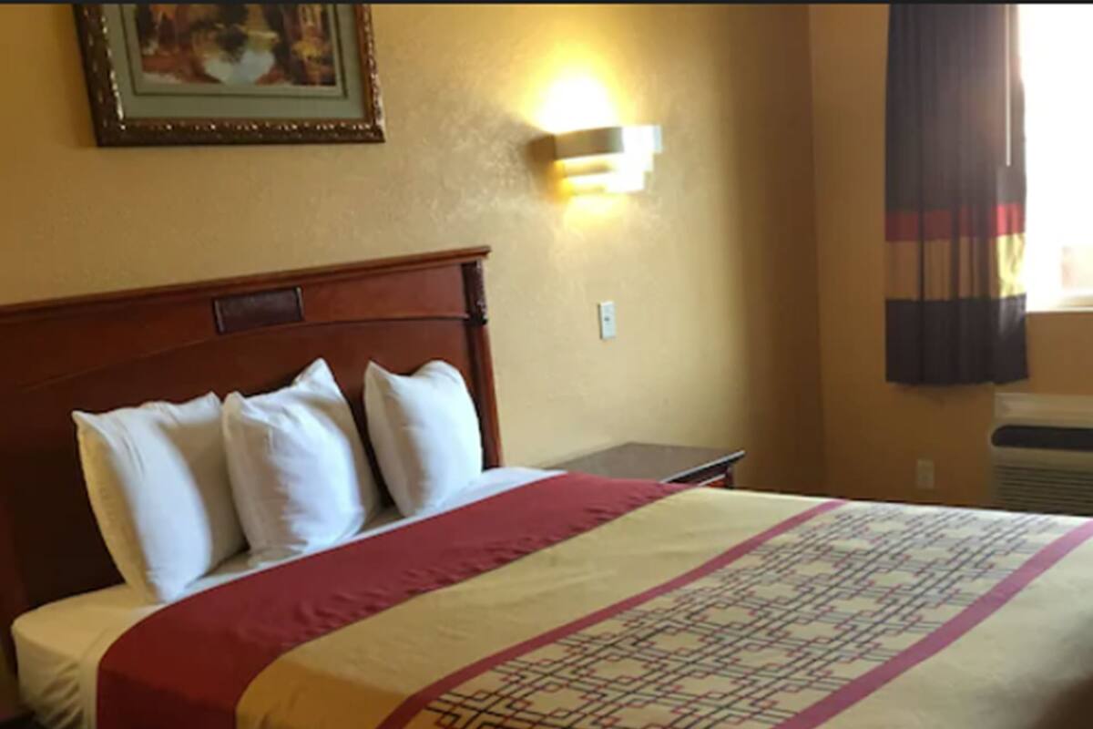 A hotel room with a wooden headboard, a bed with a patterned bedspread, a wall-mounted light, and a framed picture above the bed.