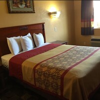 Una habitación de hotel con cabecera de madera, cama con colcha estampada, lámpara de pared e imagen enmarcada sobre la cama.