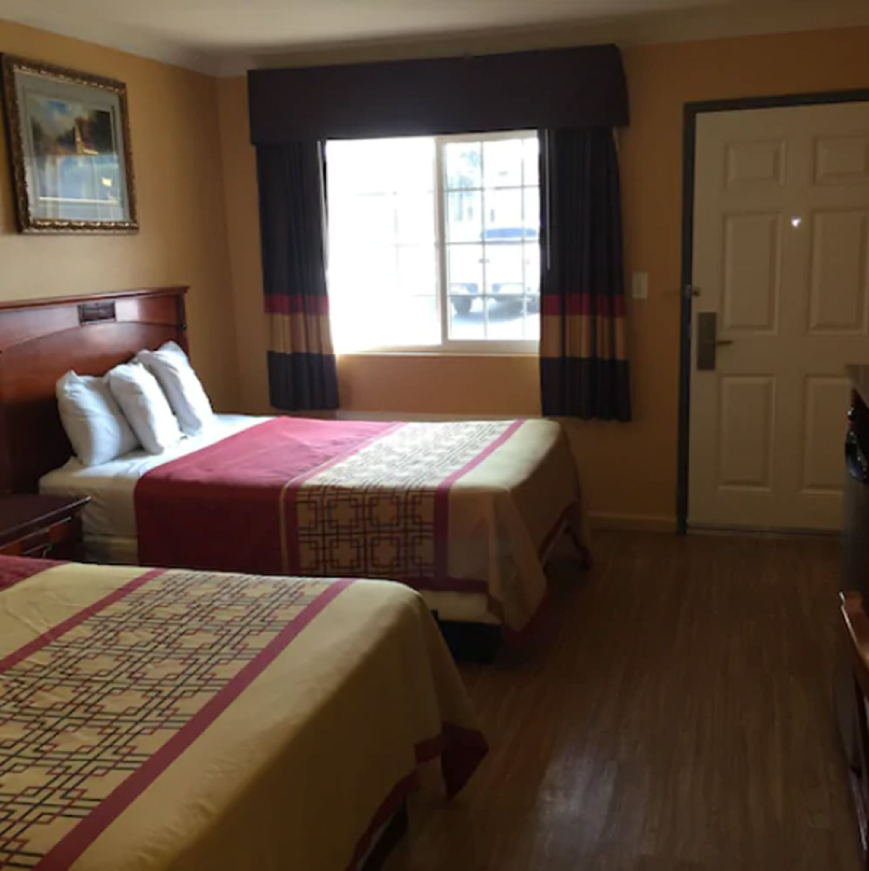 Una camera d'albergo con due letti, una testiera in legno, una finestra con tende, una porta e un quadro appeso al muro.