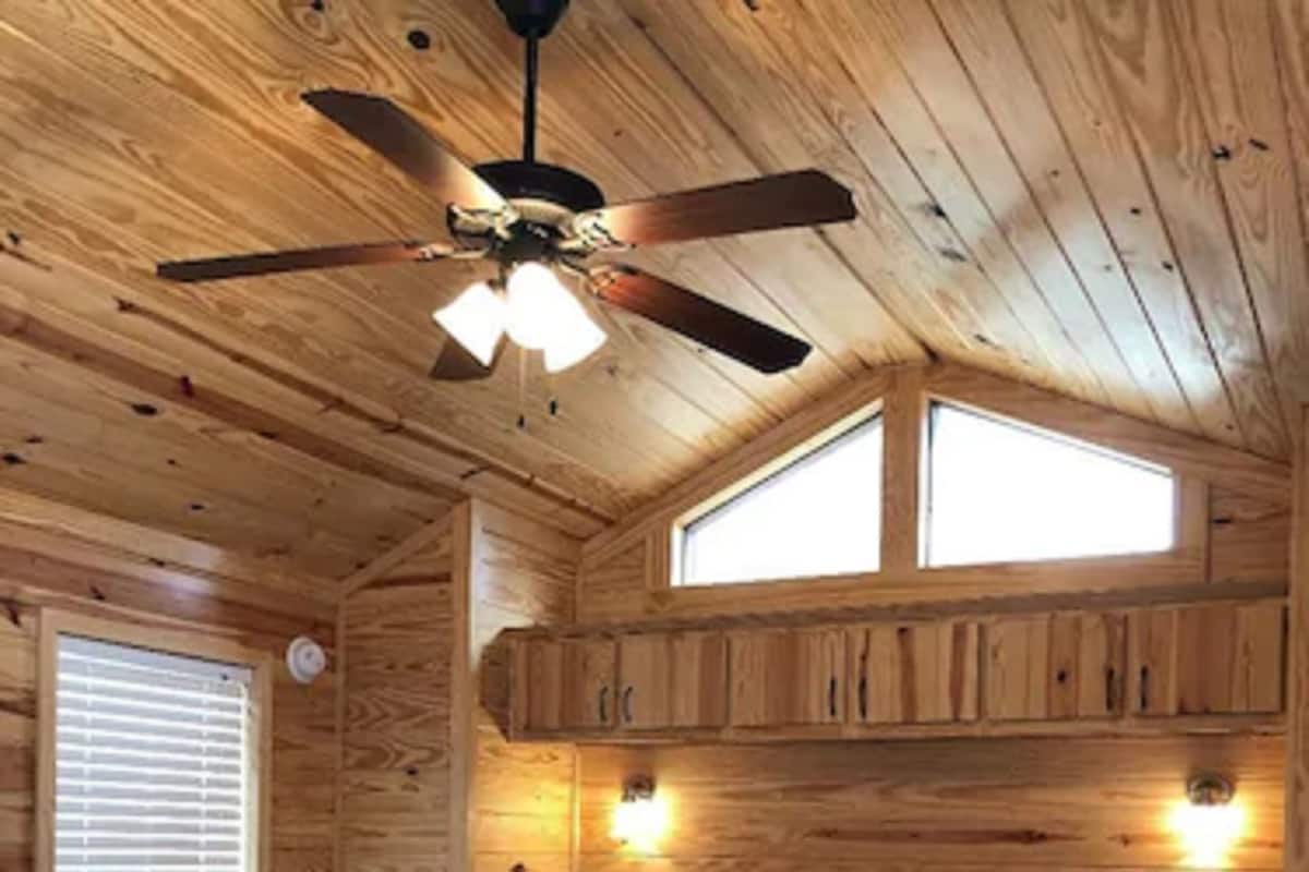 A wooden cabin bedroom with a bed, ceiling fan, two windows, and wall-mounted lights.