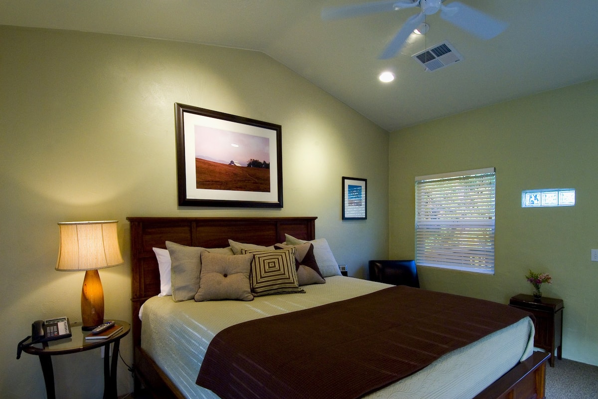 A neatly arranged bedroom with a large bed, a bedside table with a lamp, a framed picture on the wall, and a ceiling fan.