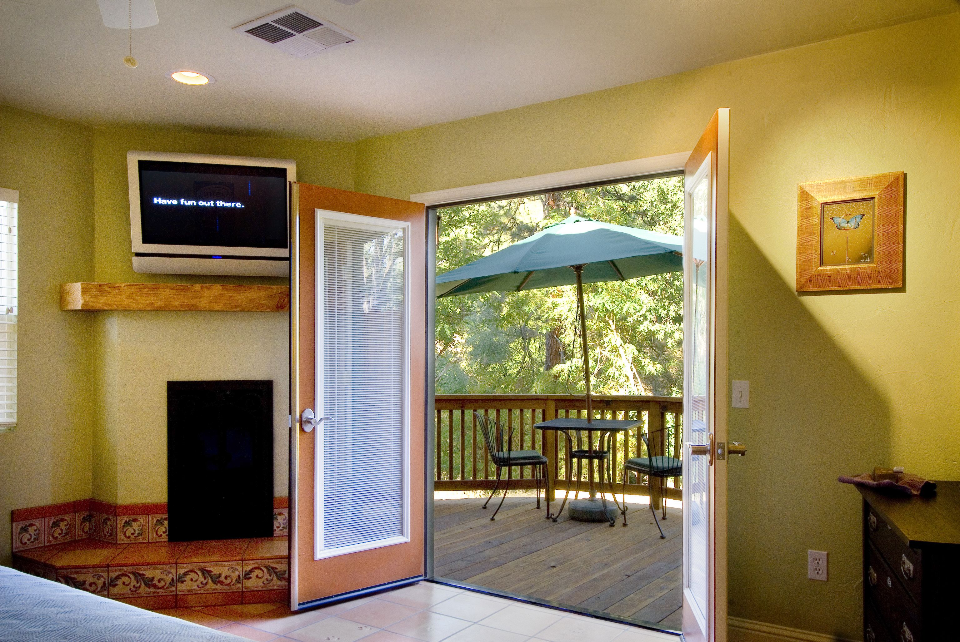 Una habitación con una televisión montada en la pared, un ventanal corredero que da a una terraza exterior con mesa y sillas, y un banco de madera.