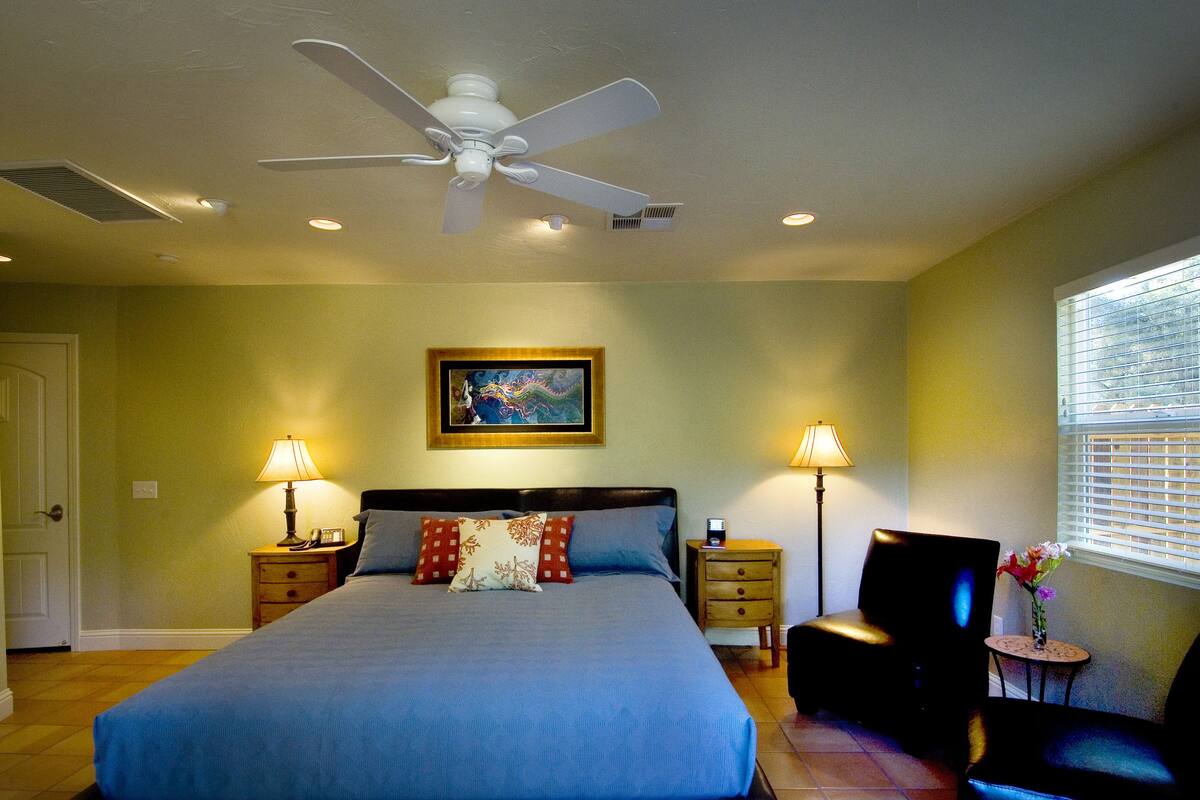 A bedroom with a bed, a ceiling fan, two bedside lamps, a chair, a small table with flowers, and a painting on the wall.