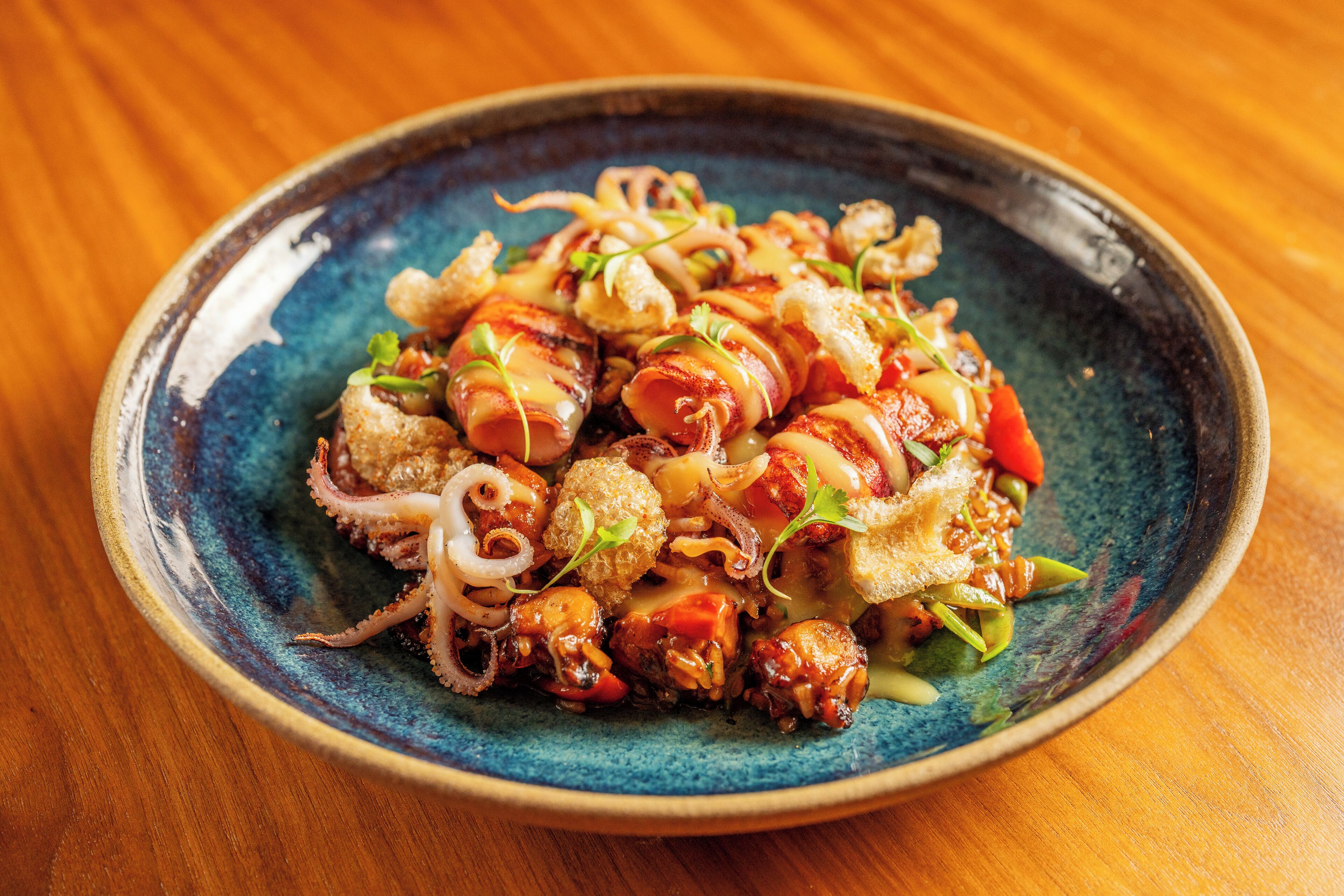 Un plato de comida con diversos ingredientes, entre ellos lo que parece ser pulpo, verduras y posiblemente algún tipo de carne, servido en un plato de cerámica azul.