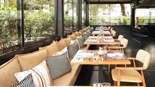 Un restaurante moderno con mesas de madera largas y asientos acolchados, grandes ventanales que ofrecen vistas a zonas verdes.