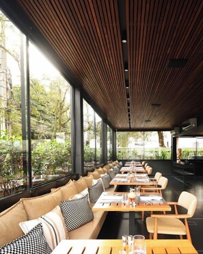 A modern restaurant with long wooden tables and cushioned seating, large windows offering a view of greenery. - Nomaa Hotel (Curitiba)