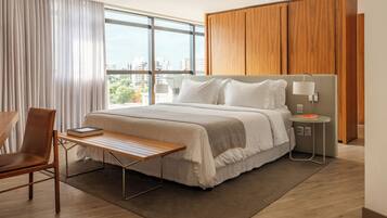 Habitación de hotel moderna con una cama grande, un armario de madera, una mesita de noche con lámpara y vistas a la ciudad a través de amplios ventanales.